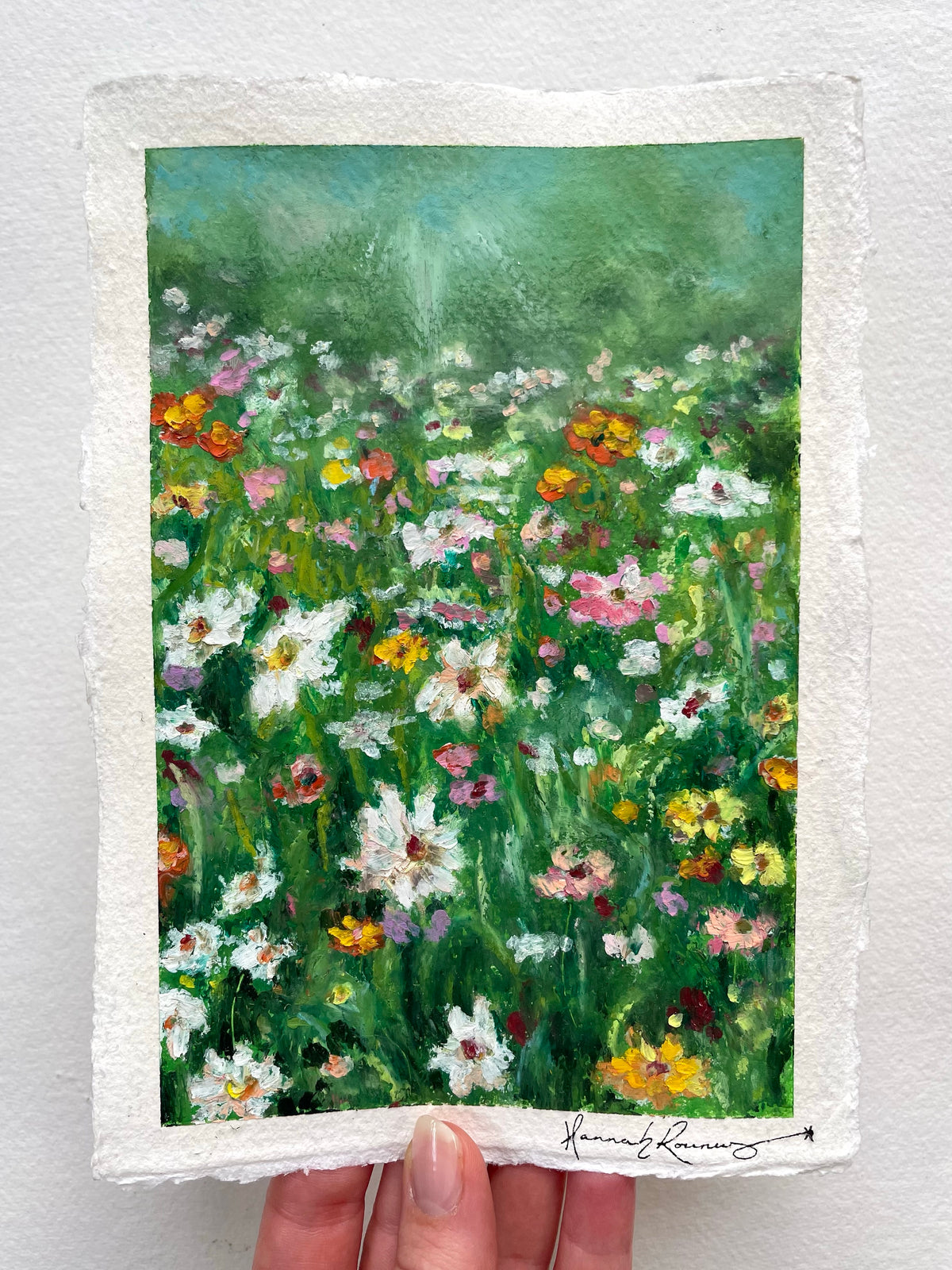 An oil pastel painting on paper depicting a vibrant wildflower meadow. The painting features green foliage and numerous white, pink, yellow, and orange flowers, with a textured white border and a signature at the bottom.