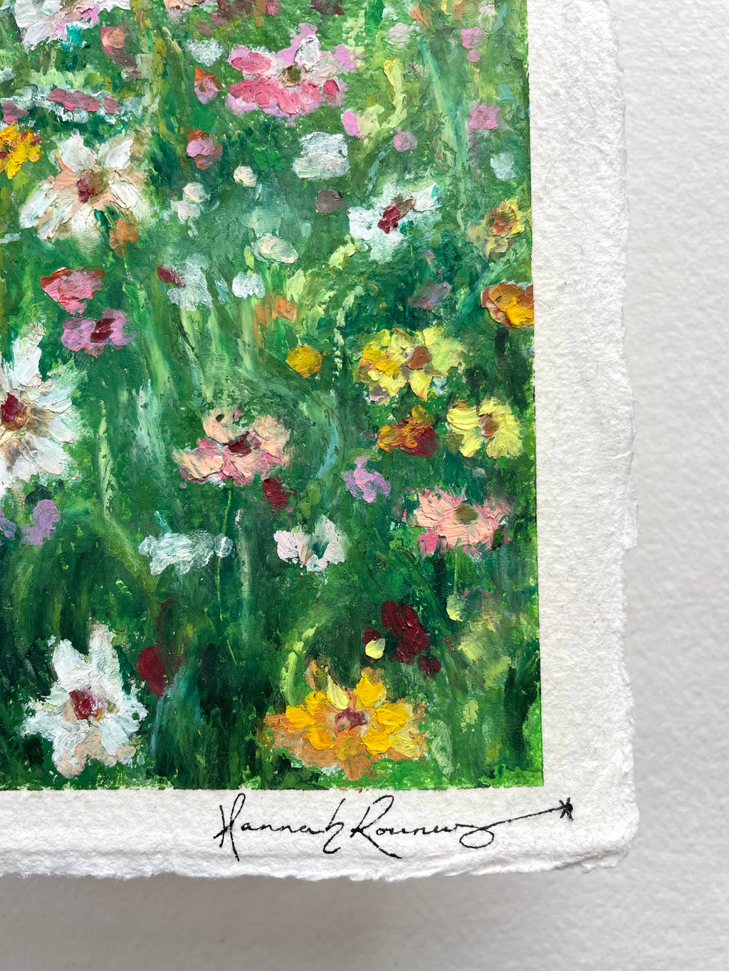 A close-up of a textured painting featuring an impressionistic wildflower meadow with green foliage and colourful blossoms. The painting has a white paper border with the artist signature at the bottom.
