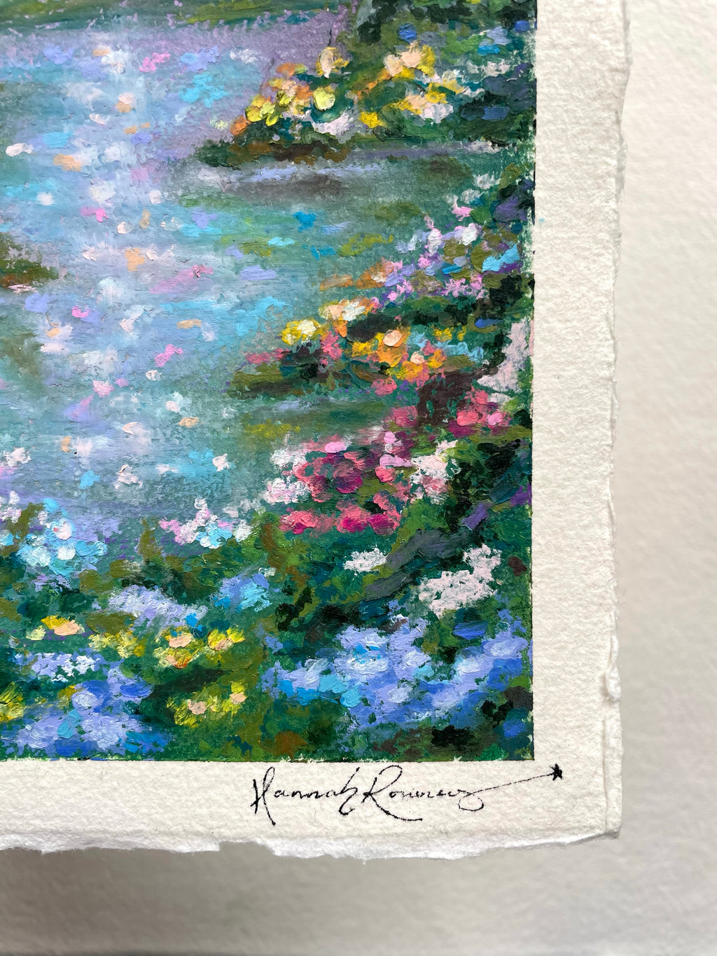 Close-up of a textured oil pastel painting on white paper, showing a forest scene with blue water, green foliage, and colourful flowers. The artist's signature is visible at the bottom.