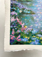 A close-up of a textured oil pastel painting on handmade paper, depicting a lush forest scene with pink, purple, blue, and green flowers against a soft river background.