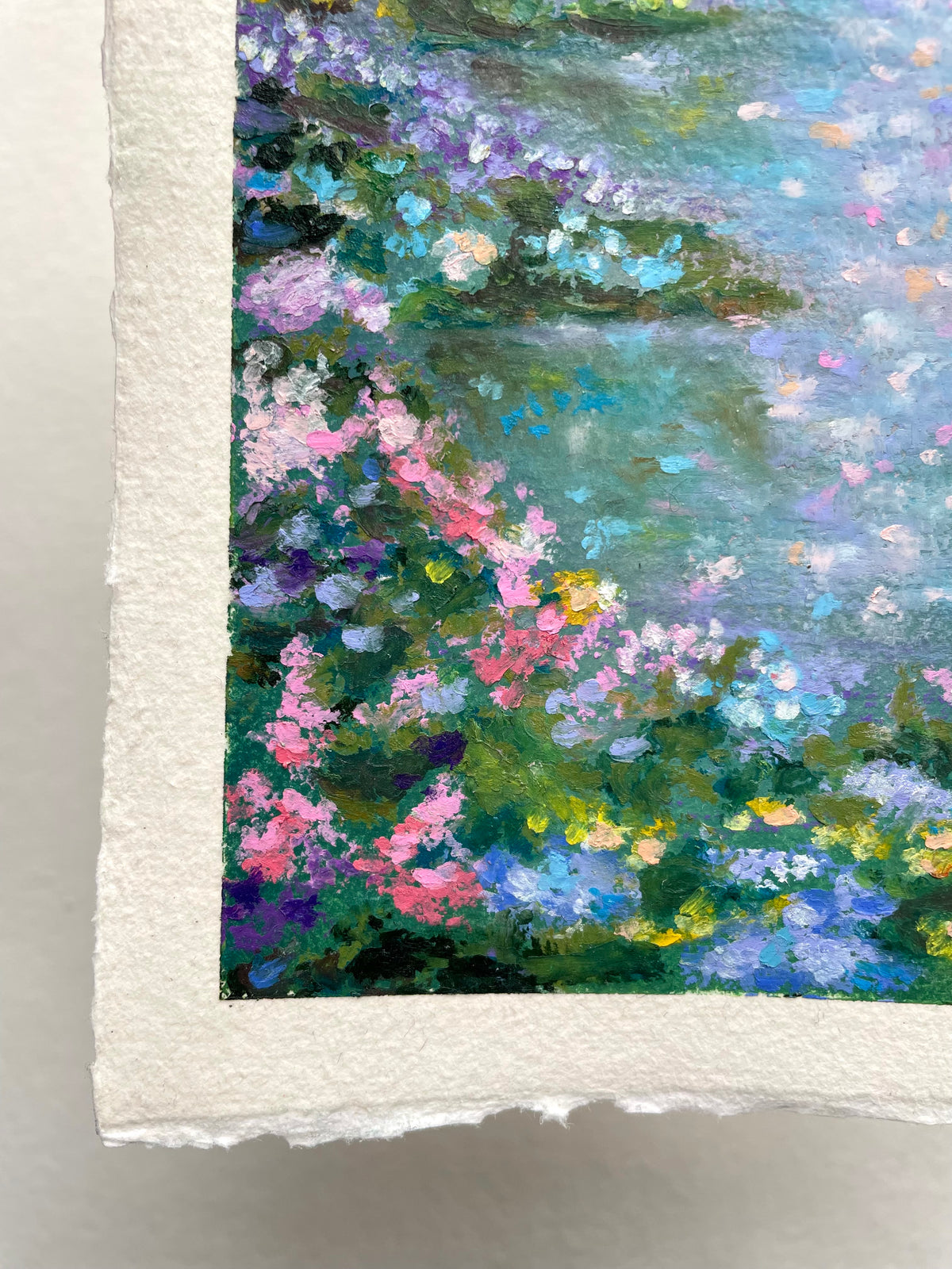 A close-up of a textured oil pastel painting on handmade paper, depicting a lush forest scene with pink, purple, blue, and green flowers against a soft river background.