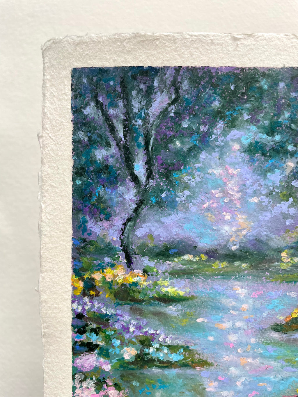A close-up of a textured oil pastel painting on paper, depicting a serene landscape with trees, flowers, and reflective water in shades of blue, purple, green, and yellow.