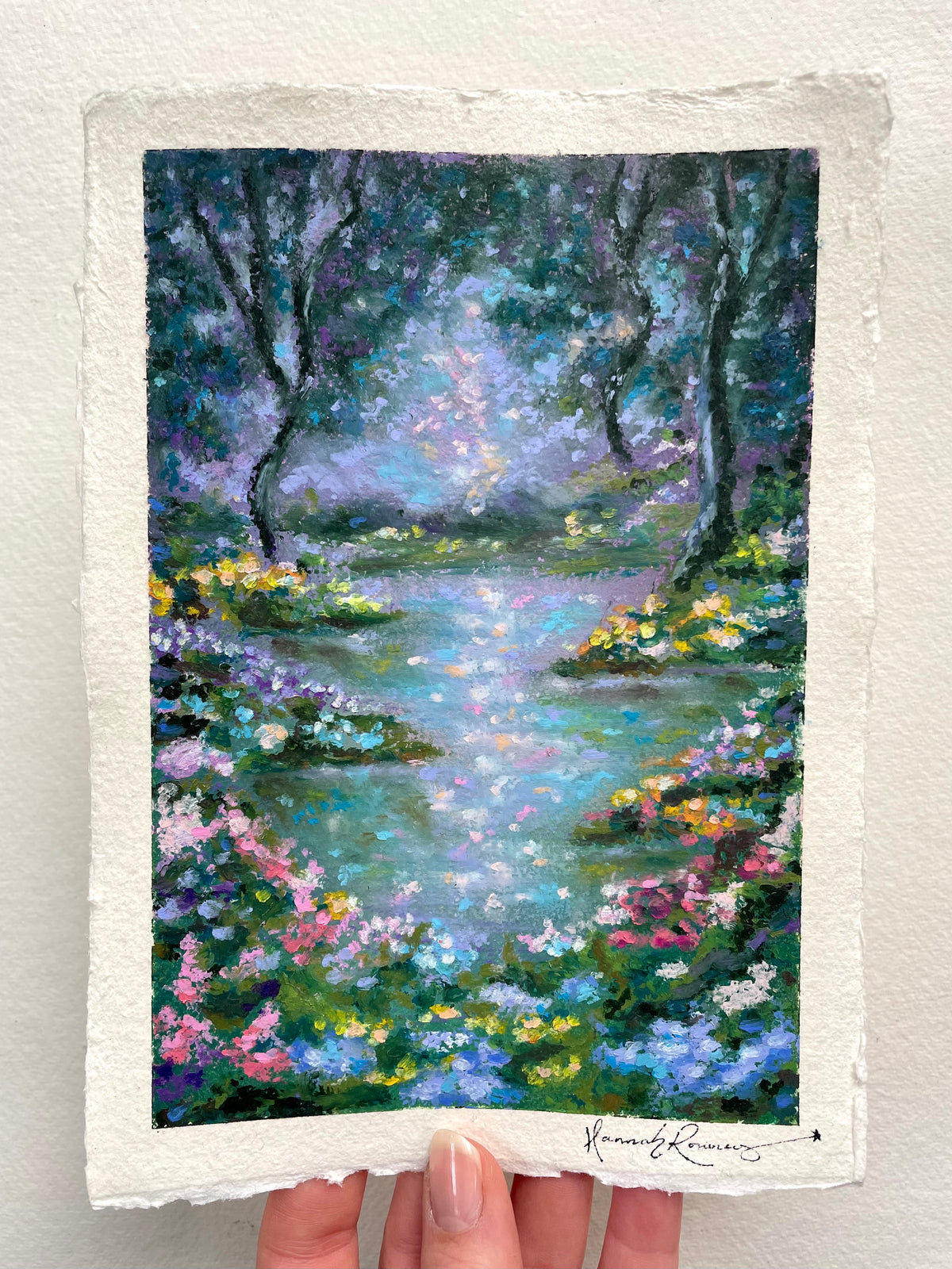 An oil pastel painting of a forest stream surrounded by colourful wildflowers, with a textured off-white paper border and a visible artist signature at the bottom.