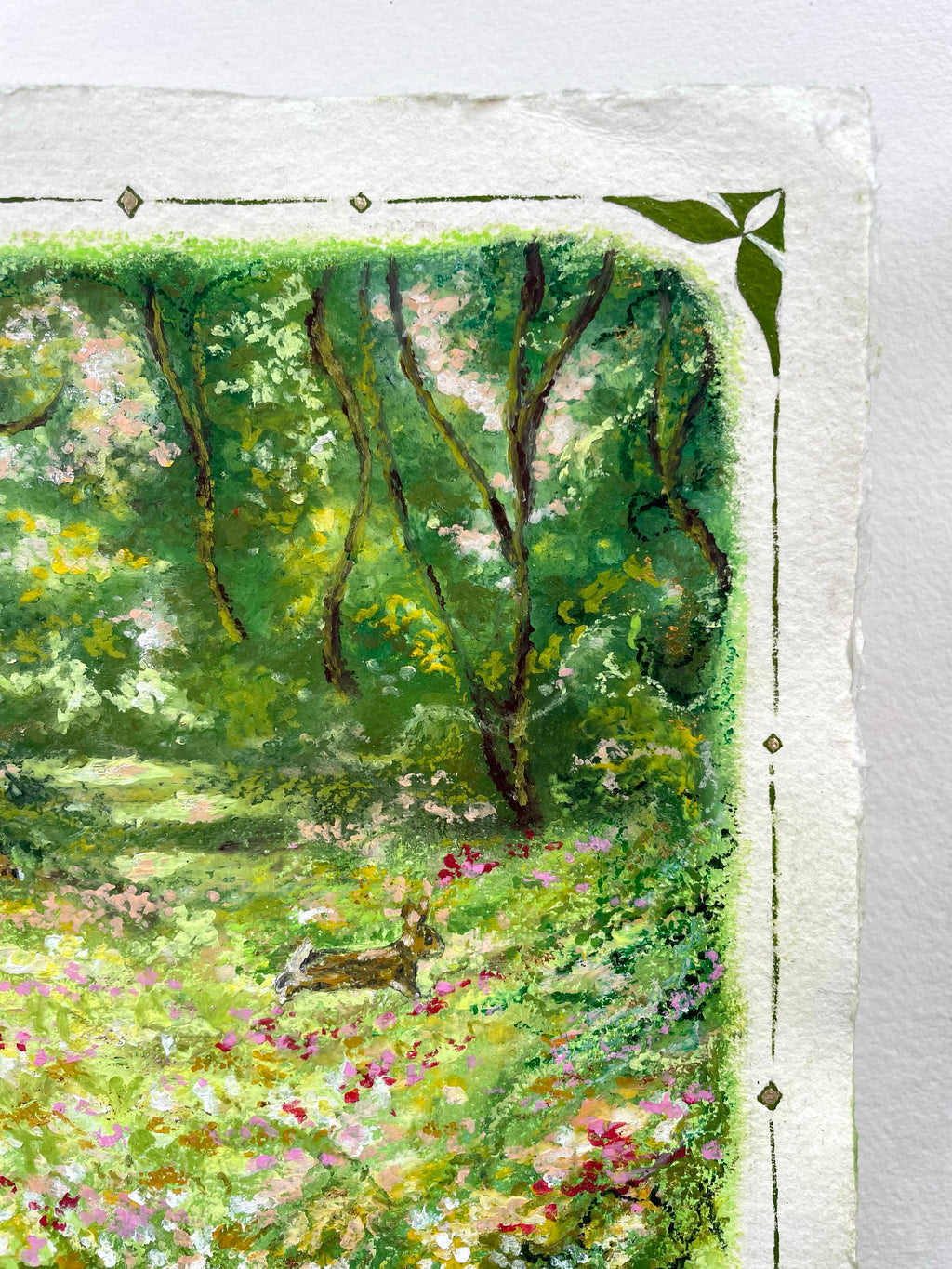 A close-up of an oil pastel painting on textured paper, depicting a lush green forest scene with trees, a rabbit and wildflowers, framed by a white border with decorative corner and side motifs.