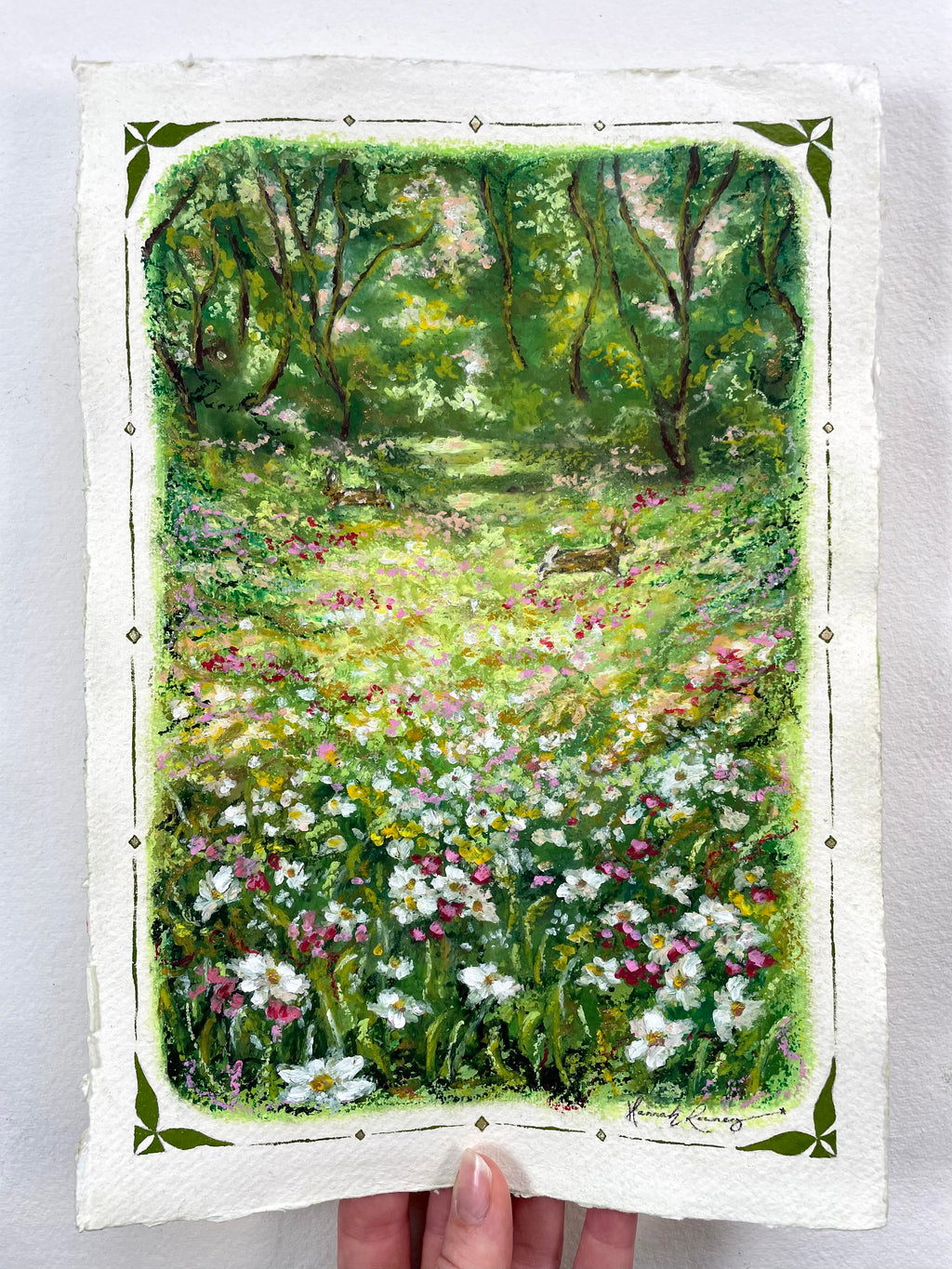 A hand holds an oil pastel painting featuring a colourful landscape of a sunlit woodland meadow filled with white, pink, and yellow wildflowers, with a green forest path, trees and two rabbits in the background. The artwork has a white border with a decorative green corner pattern.