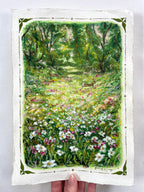 A hand holds an oil pastel painting featuring a colourful landscape of a sunlit woodland meadow filled with white, pink, and yellow wildflowers, with a green forest path, trees and two rabbits in the background. The artwork has a white border with a decorative green corner pattern.