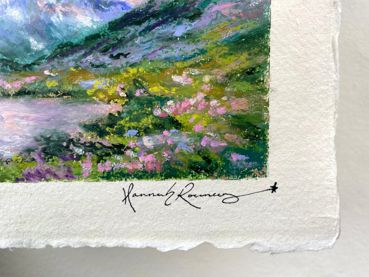 Close-up of an oil pastel landscape painting showing green and purple hills, pink flowers, and a lake, with a white textured paper border and an artist signature.