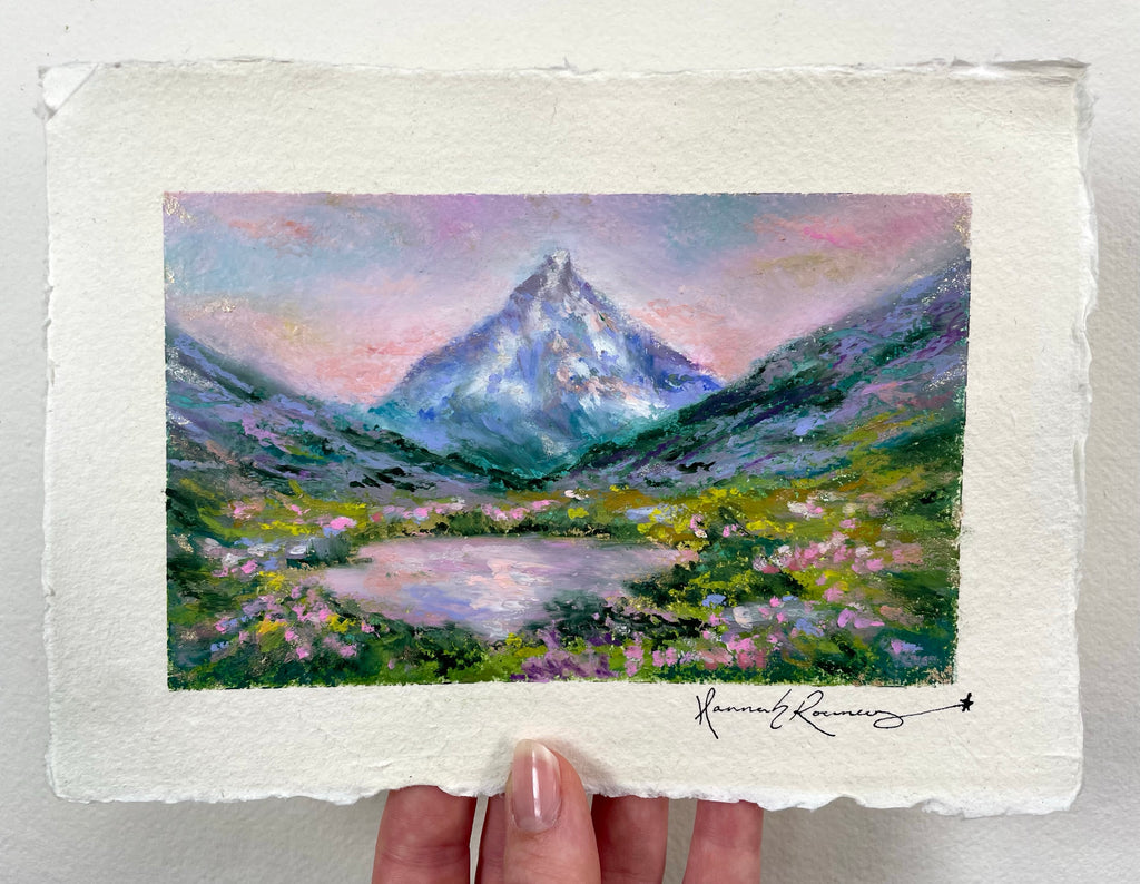 A hand holds an oil pastel painting of a mountain landscape with a lake and wildflowers. The artwork shows a snow-capped peak under a pastel pink and purple sky, with green and blue mountains and a reflective lake in the foreground.