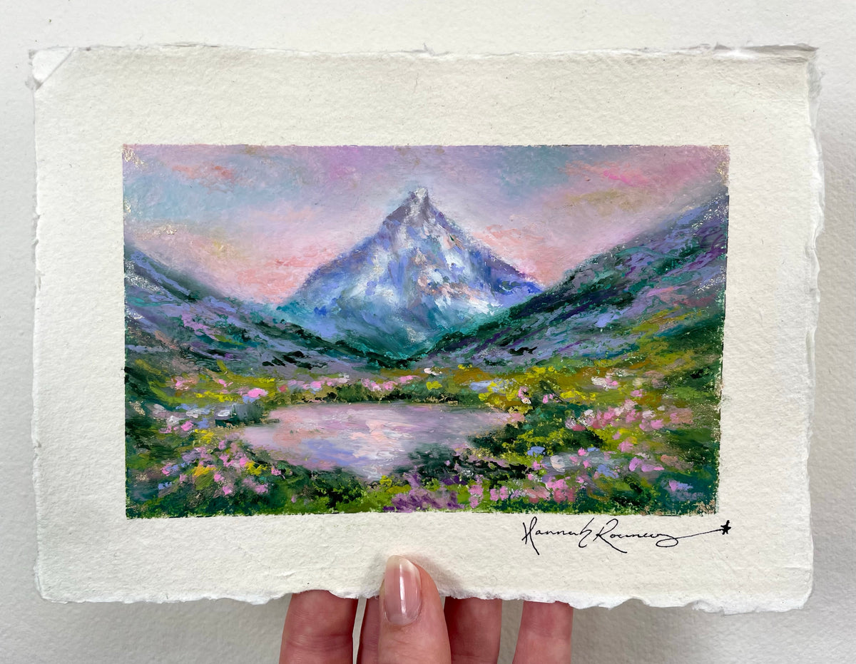 A hand holds an oil pastel painting of a mountain landscape with a lake and wildflowers. The artwork shows a snow-capped peak under a pastel pink and purple sky, with green and blue mountains and a reflective lake in the foreground.
