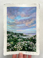 A hand holds an oil pastel painting on paper of a coastal landscape with a dramatic blue and purple sky, green hills, and pink-and-white flowers. The artist's signature is visible in the bottom right corner.