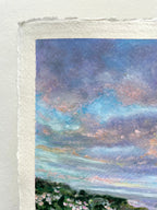 A close-up of a textured oil pastel landscape painting on paper showing a dramatic sky with swirling blue, purple, and pink clouds above a dark green landscape with small pale flowers. The painting has a rough, handmade paper border.