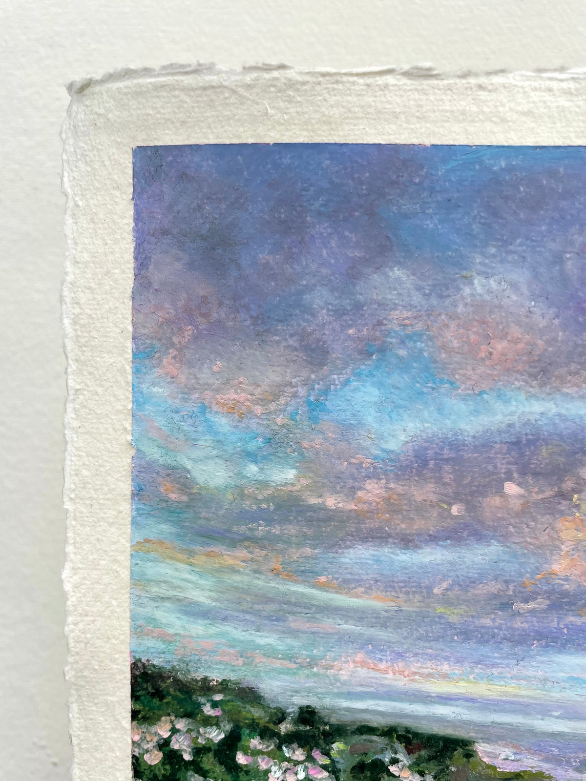 A close-up of a textured oil pastel landscape painting on paper showing a dramatic sky with swirling blue, purple, and pink clouds above a dark green landscape with small pale flowers. The painting has a rough, handmade paper border.