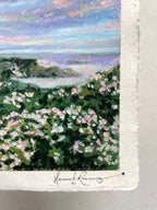 A close-up of an oil pastel painting on paper showing a coastal landscape with green hills covered in pink and white flowers under a pastel sky, with a visible artist signature at the bottom.