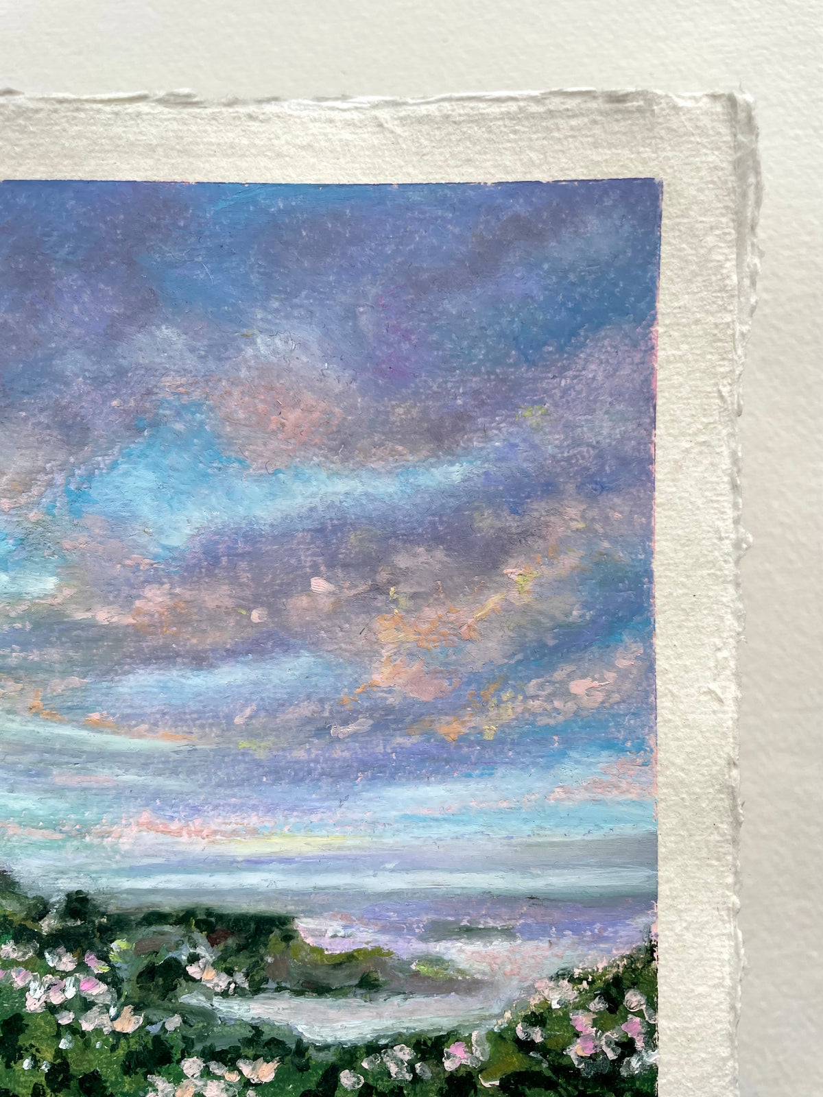 A close-up of an oil pastel landscape painting on paper featuring a seascape with a dramatic blue and purple sky, soft pink clouds, and green coastal foliage with small pink flowers.