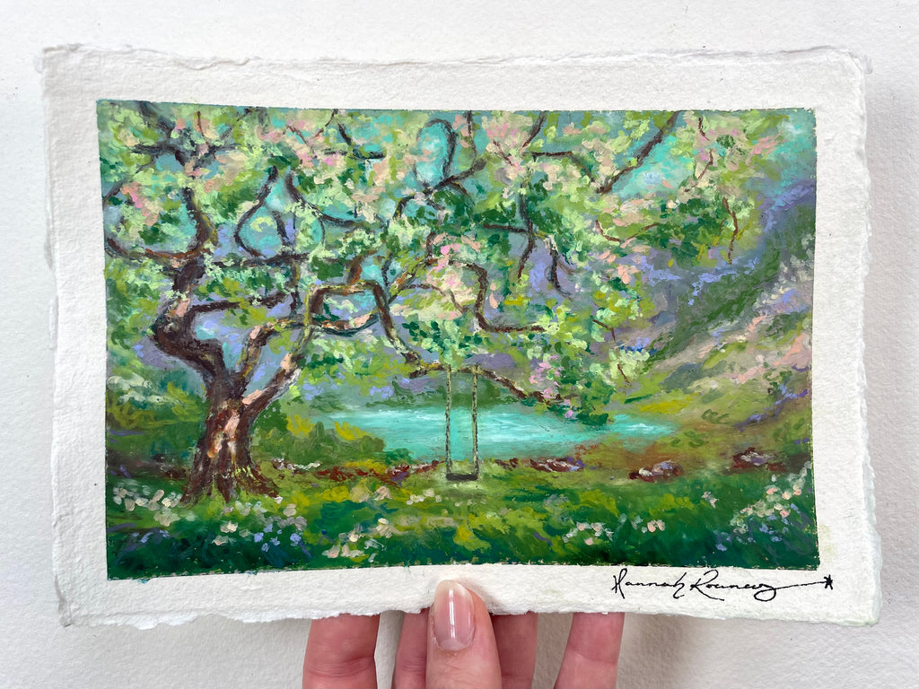 An oil pastel painting on textured white paper, depicting a lush green landscape with a large tree, a swing, and a turquoise lake. The artist’s signature is in the bottom right corner.