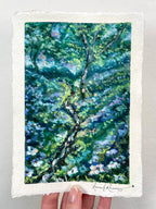 A hand holds an oil pastel painting of a tree with dense green, blue, and purple foliage and white flowers.