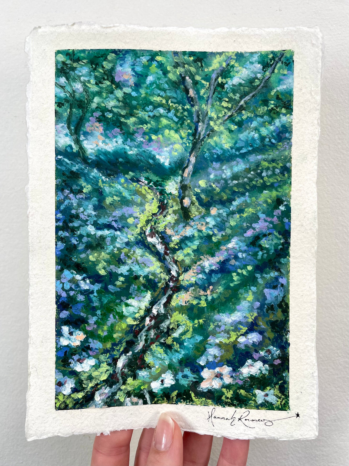 A hand holds an oil pastel painting of a tree with dense green, blue, and purple foliage and white flowers.