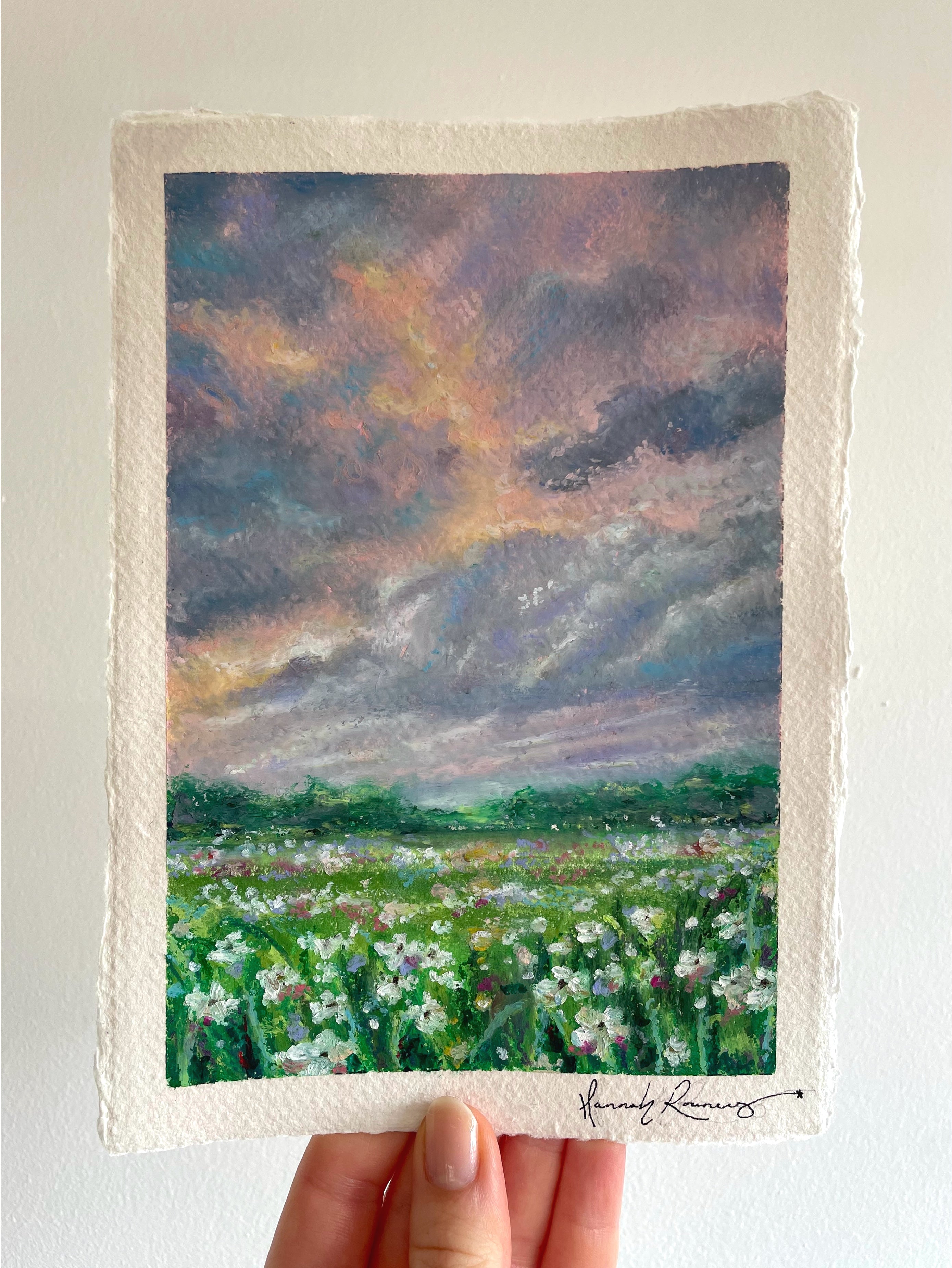 Original whimsical oil pastel landscape painting held in hand, featuring glowing storm-lit sky, colourful wildflower meadow, and magical light breaking through dramatic clouds.