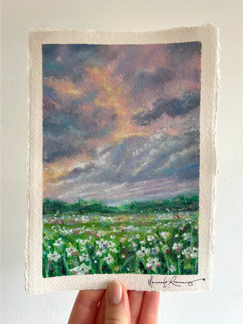 Original whimsical oil pastel landscape painting held in hand, featuring glowing storm-lit sky, colourful wildflower meadow, and magical light breaking through dramatic clouds.