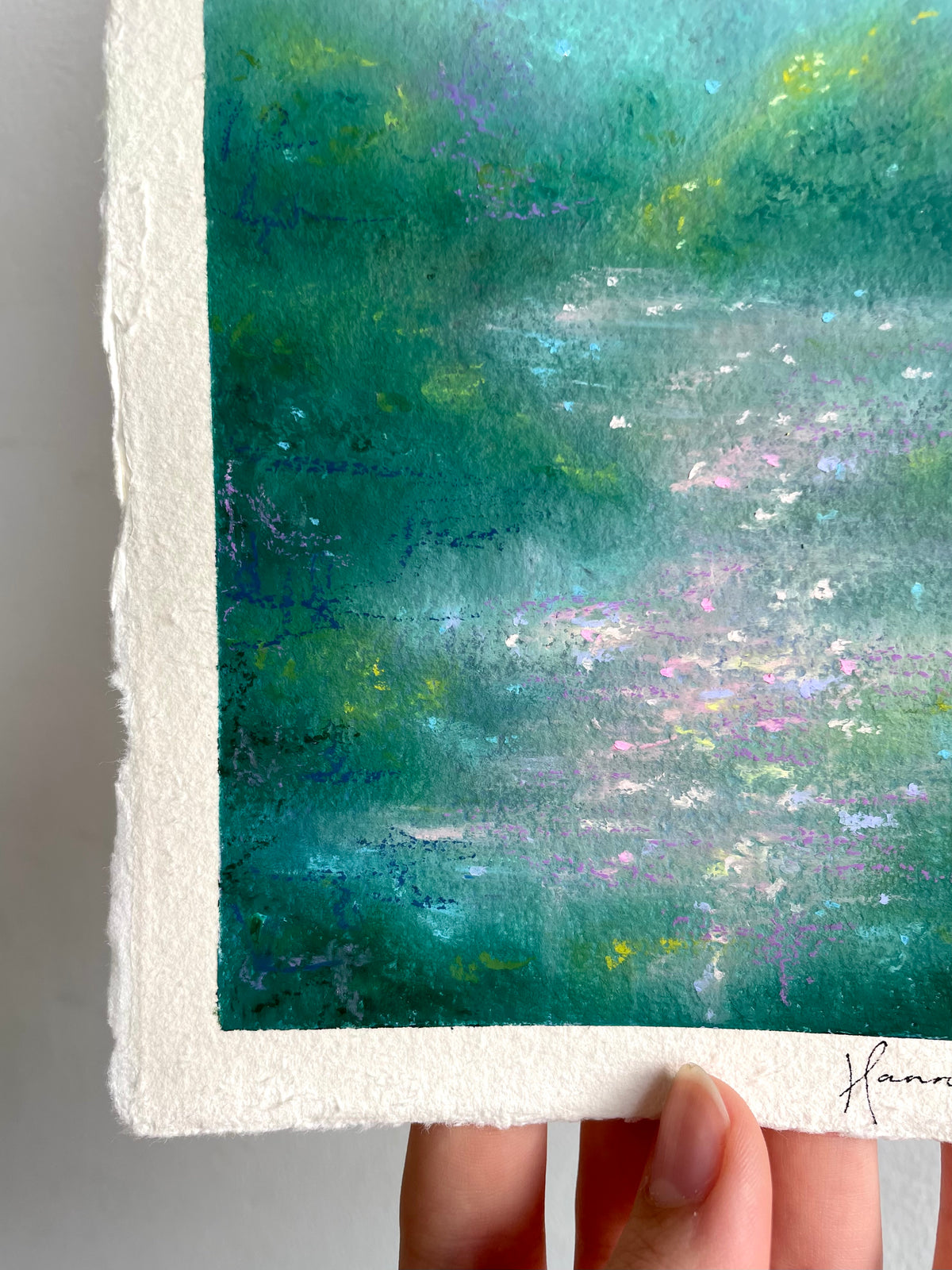 A close-up of an oil pastel painting on handmade paper, held by a hand. The artwork features a serene landscape with teal, green, and blue tones, white highlights, and subtle purple and yellow accents.