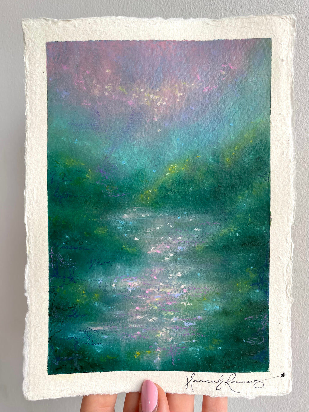 An impressionistic landscape painting on textured white paper, held by a hand. The artwork depicts a serene, misty scene with deep green and teal water, foliage, soft purple and lavender light, and a shimmering sky.