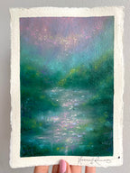 An impressionistic landscape painting on textured white paper, held by a hand. The artwork depicts a serene, misty scene with deep green and teal water, foliage, soft purple and lavender light, and a shimmering sky.