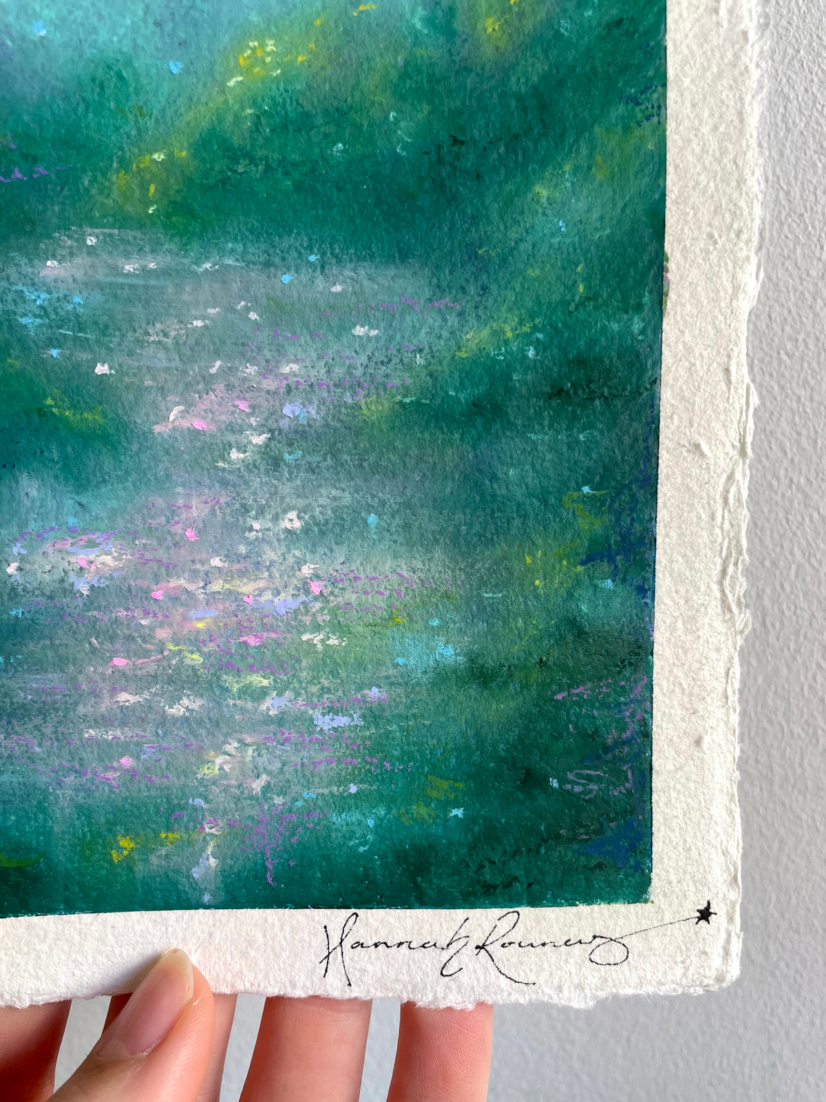 A close-up of a magical oil pastel painting on handmade paper, held by a hand. The artwork features a blend of deep teal, green, and blue tones with soft lavender and white highlights, finished with a white border and an artist signature at the bottom.