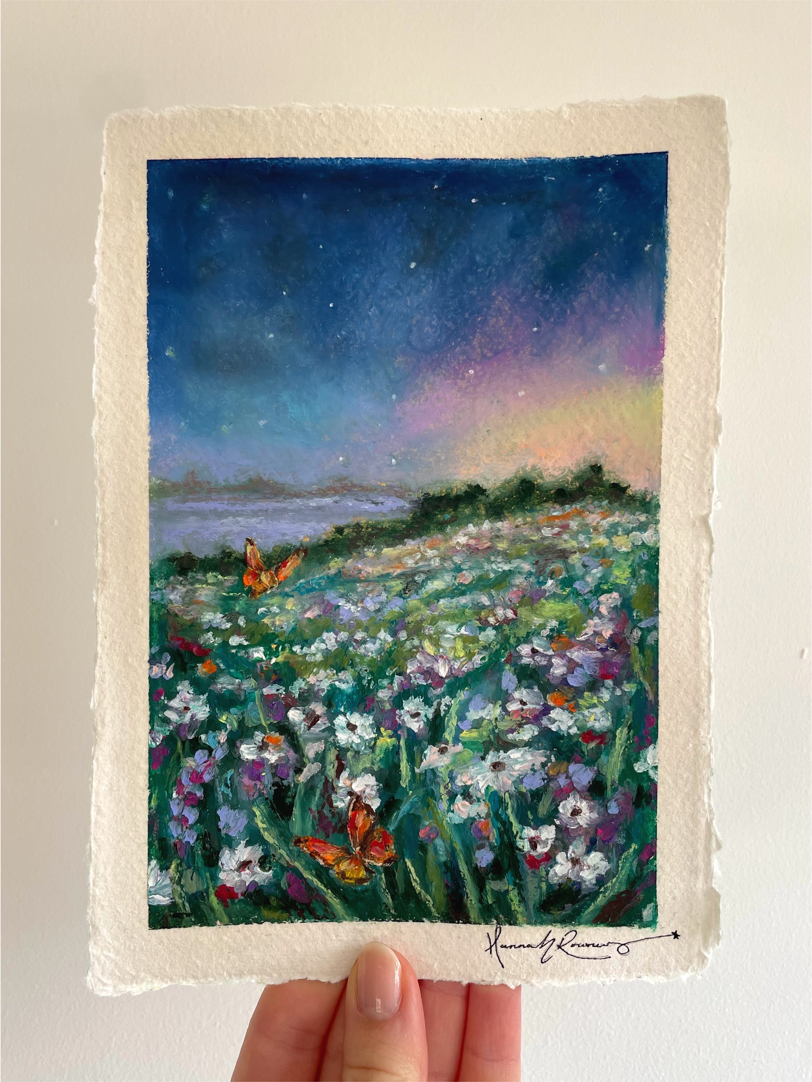 Original mystical oil pastel landscape painting held in hand, featuring glowing orange butterflies, dreamy midnight sky, distant lake, and richly textured wildflower scenery.