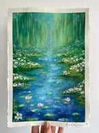 Original oil pastel impressionist painting ‘Lighter Than The Lilies’ held against a white wall, featuring a magical pond landscape with water lilies, lily pads, wildflowers, soft weeping willow trees, sparkling sunlight and dreamy turquoise blue and green tones.