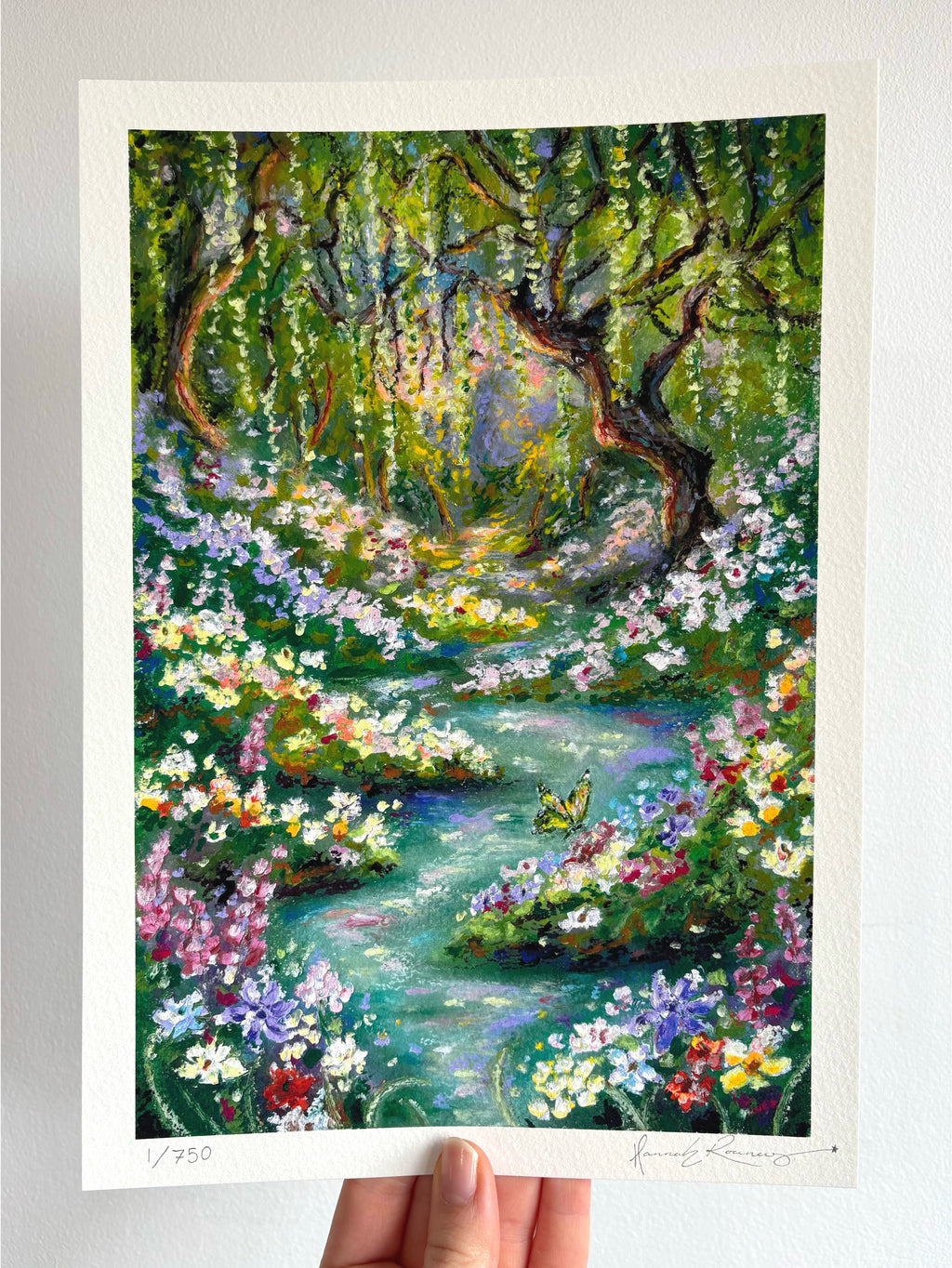 Limited edition giclée fine art print of whimsical oil pastel landscape “Looking for Wonderland,” featuring enchanted river, butterfly, wildflowers, and glowing weeping willow held against a white wall.