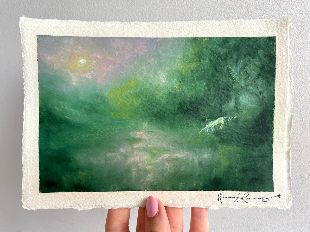 A hand holds a landscape oil pastel painting of a misty green forest with a small white unicorn and a glowing sun, featuring a textured off-white paper border and an artist signature in the bottom right corner.