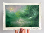 A hand holds a landscape oil pastel painting of a misty green forest with a small white unicorn and a glowing sun, featuring a textured off-white paper border and an artist signature in the bottom right corner.