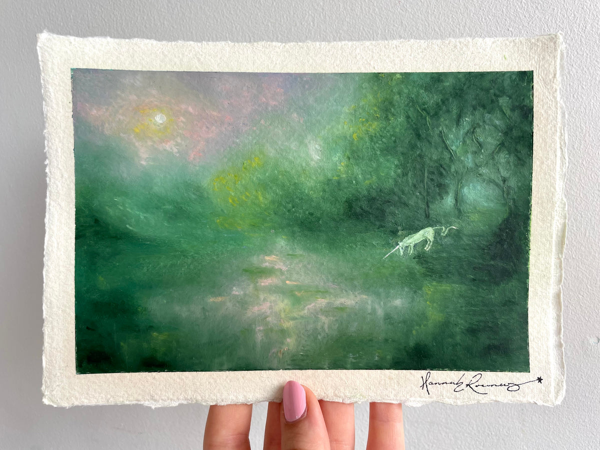 A hand holds a landscape oil pastel painting of a misty green forest with a small white unicorn and a glowing sun, featuring a textured off-white paper border and an artist signature in the bottom right corner.