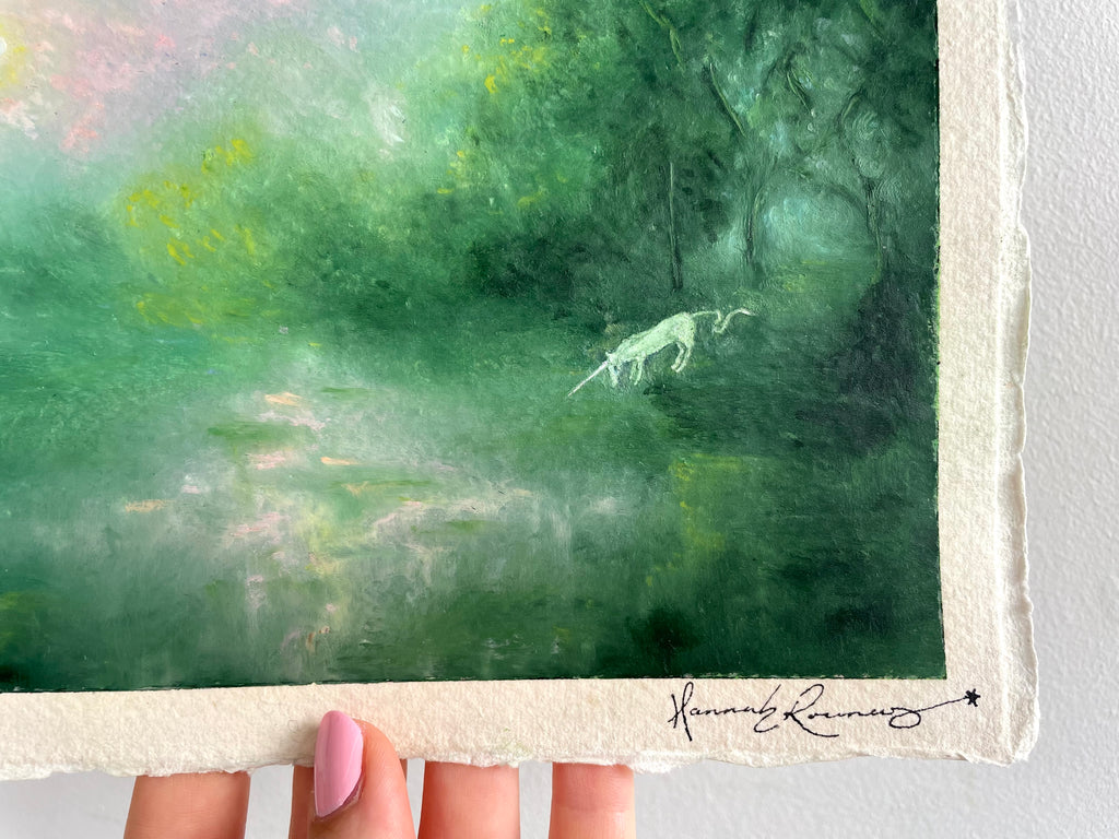 A close-up of a textured paper painting featuring a green forest scene with a small white unicorn, held by a hand. The artist's signature 'Hannah Rowney' is visible in black ink at the bottom right.