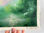 A close-up of a textured paper painting featuring a green forest scene with a small white unicorn, held by a hand. The artist's signature 'Hannah Rowney' is visible in black ink at the bottom right.