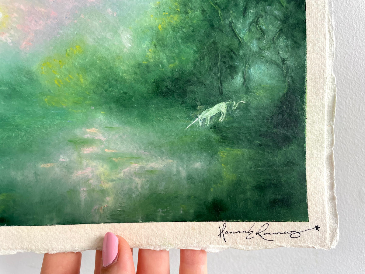 A close-up of a textured paper painting featuring a green forest scene with a small white unicorn, held by a hand. The artist's signature 'Hannah Rowney' is visible in black ink at the bottom right.