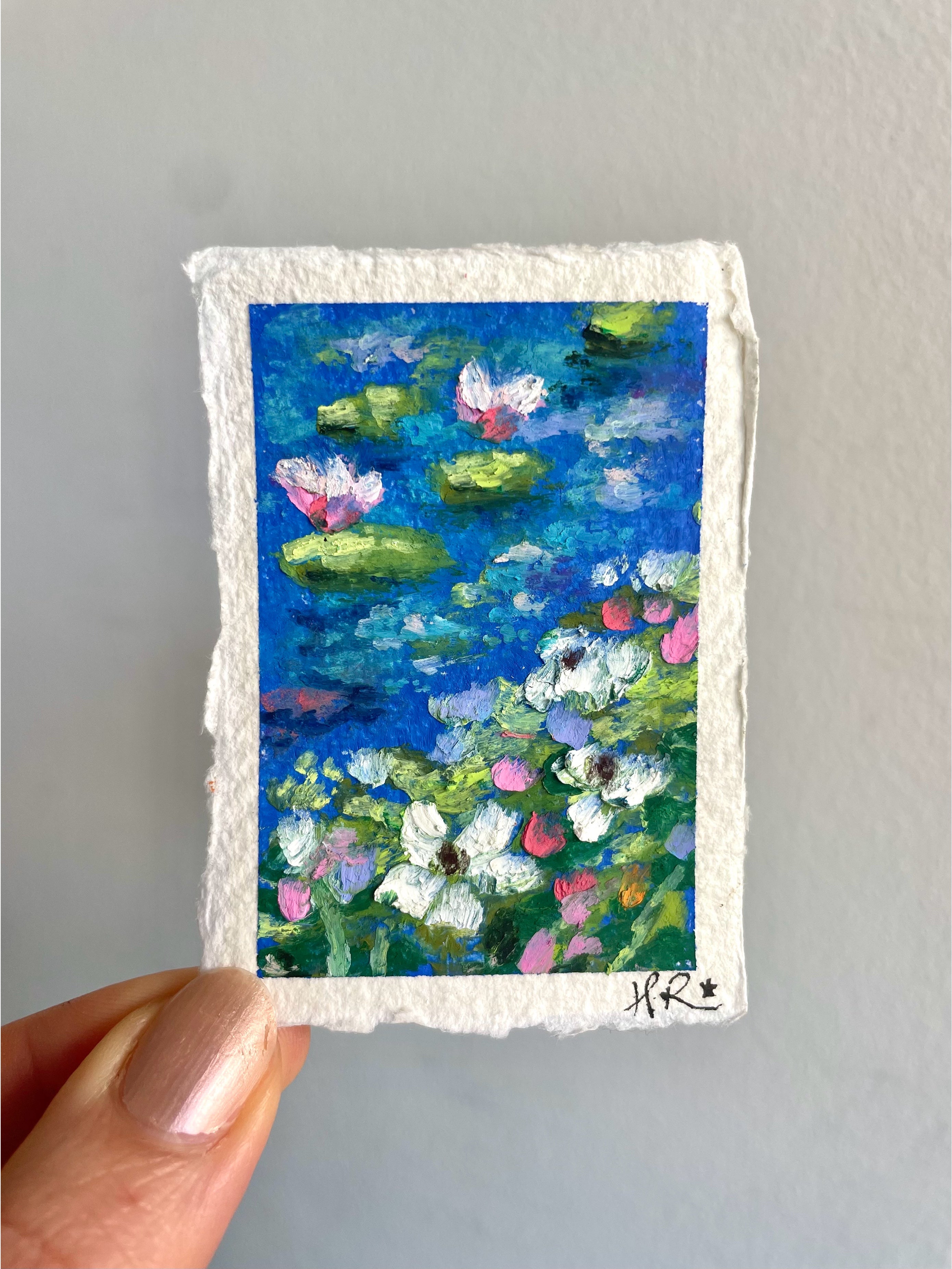 Whimsical mini oil pastel painting of deep blue pond with lily pads, water lilies, and grassy wildflower bank in a dreamy natural landscape.