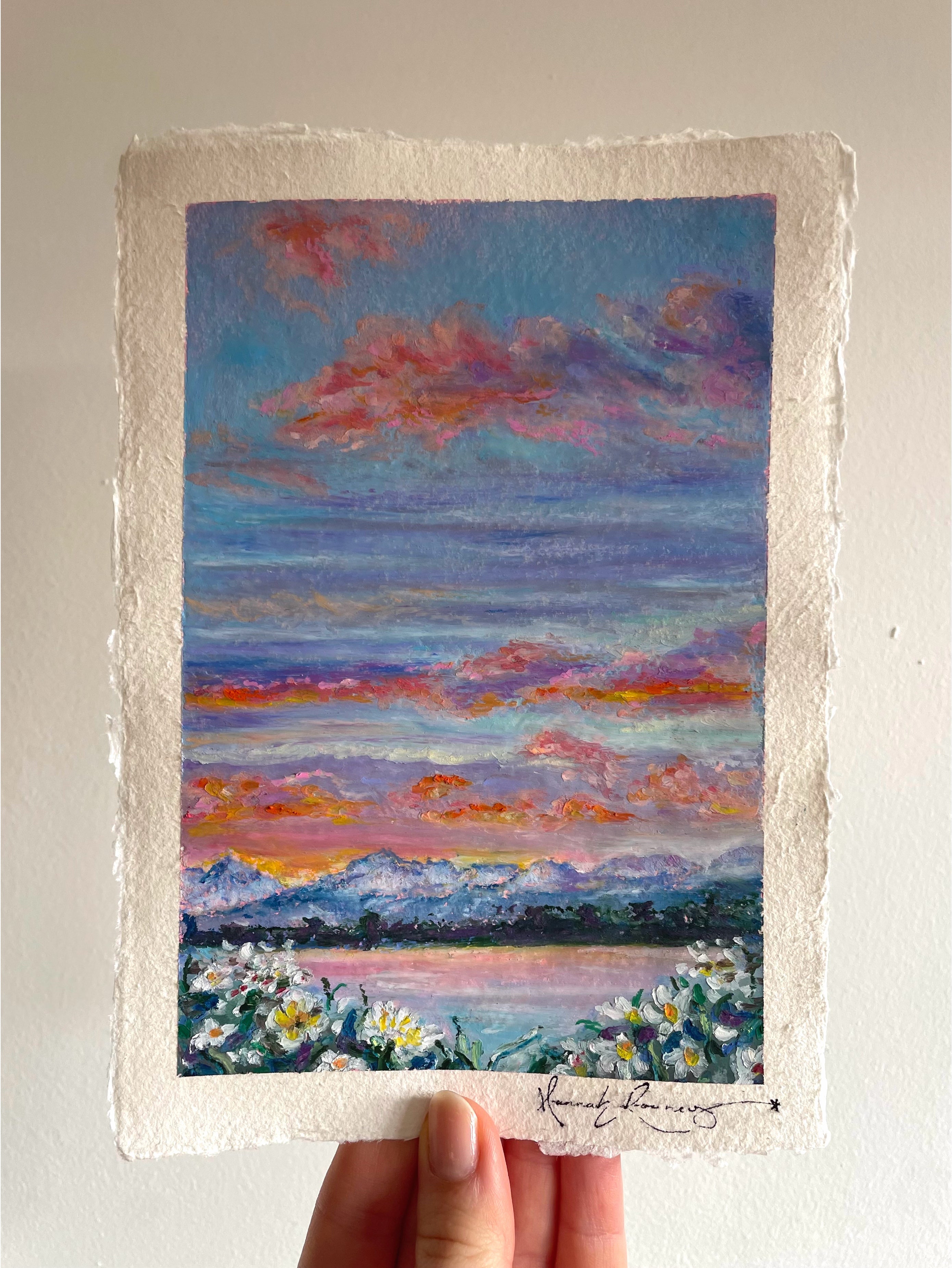 Original oil pastel mountain landscape painting held in hand, featuring glowing sunrise, calm lake, colourful wildflowers, and vibrant peaceful scenery.