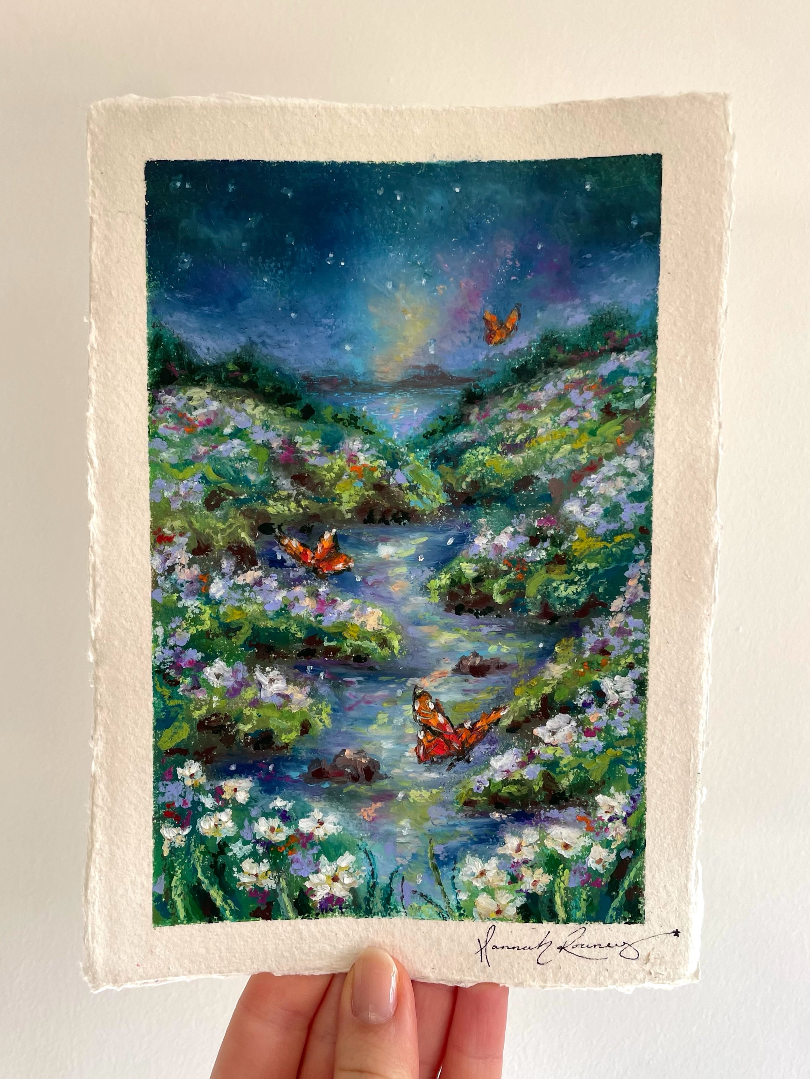 Original magical nighttime oil pastel landscape painting held in hand, featuring glowing butterflies, wildflowers, river, distant ocean, and dreamy moonlit atmosphere.