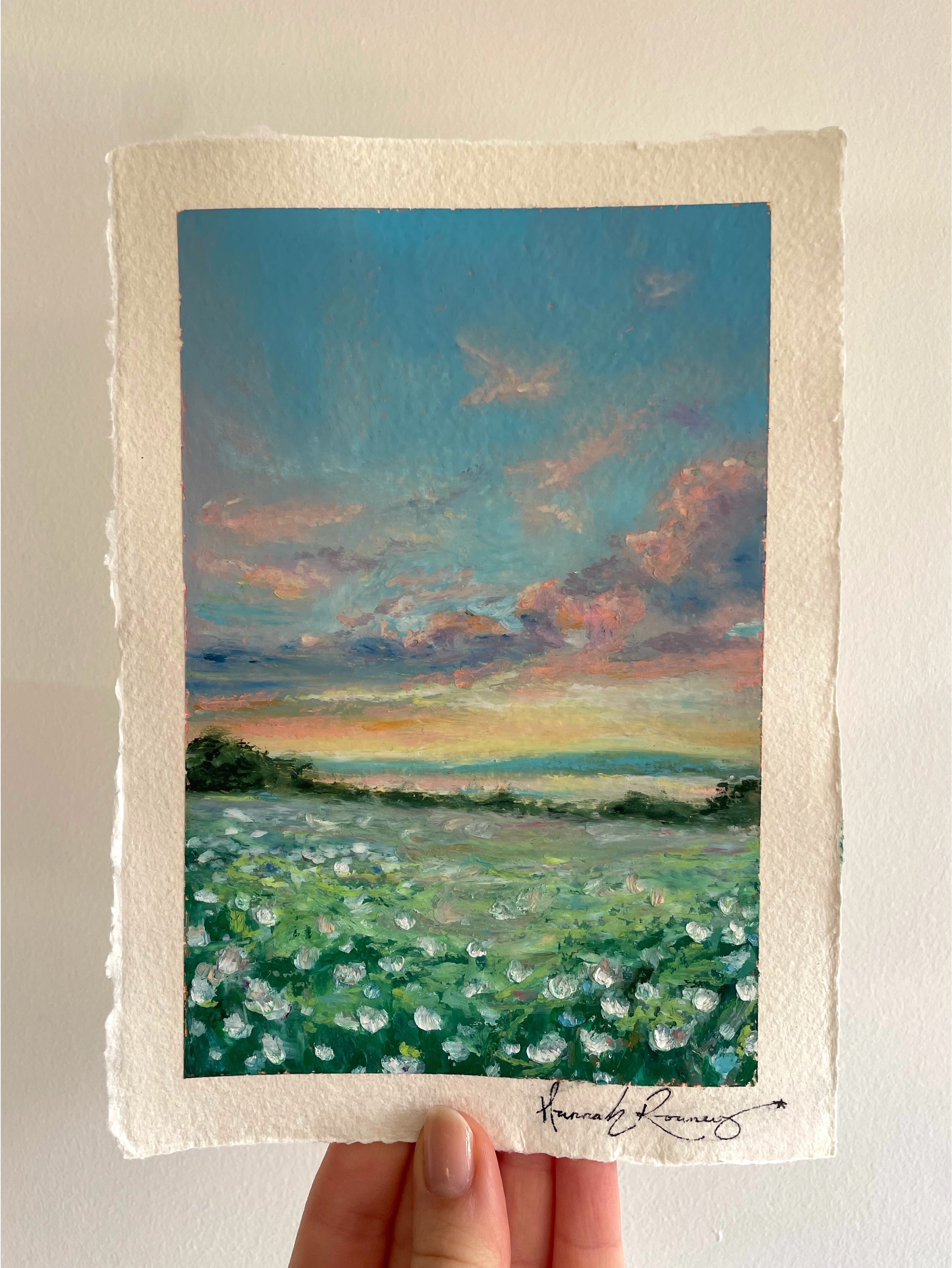 Original whimsical oil pastel sunset landscape painting held in hand, featuring glowing sky, colourful wildflowers, and dreamy nostalgic atmosphere in rich blue, pink, green, and orange tones.