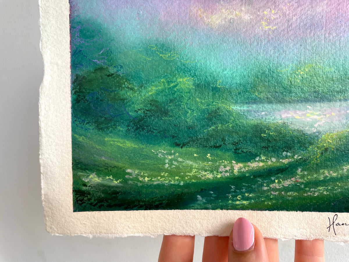 A close-up of an oil pastel landscape painting held by a hand. The painting features rolling green hills, a reflective body of water, and a pastel sky with purple, pink, and blue hues.