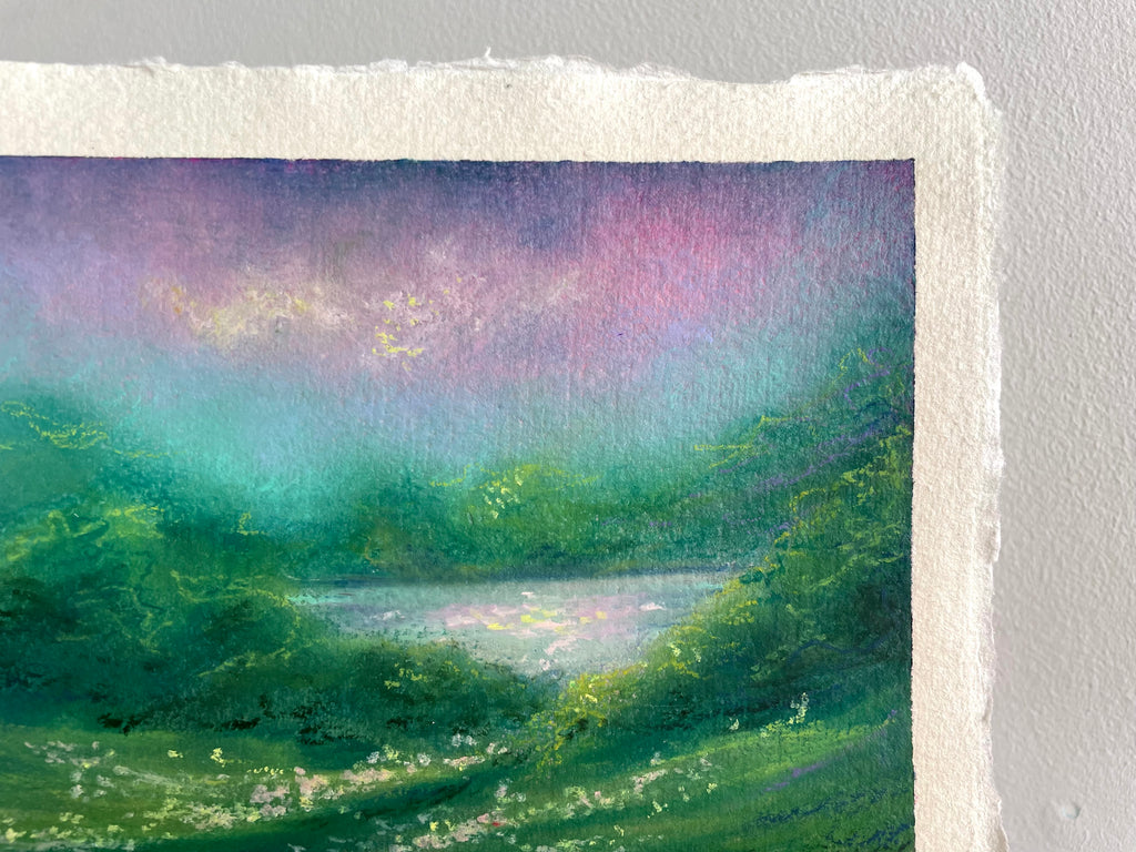 A close-up of an oil pastel landscape painting on textured paper, showing a green field, a reflective lake, and a purple and teal sky, mounted against a light gray wall.