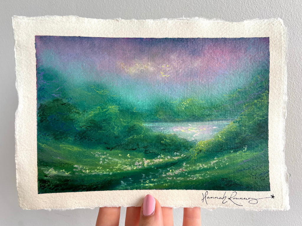 A hand holds a rectangular piece of textured handmade paper featuring an oil pastel painting of a green valley with a lake under a purple and teal sky. The artist's signature, 'Hannah Rowney', is visible in the bottom right corner.