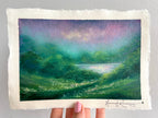 A hand holds a rectangular piece of textured handmade paper featuring an oil pastel painting of a green valley with a lake under a purple and teal sky. The artist's signature, 'Hannah Rowney', is visible in the bottom right corner.