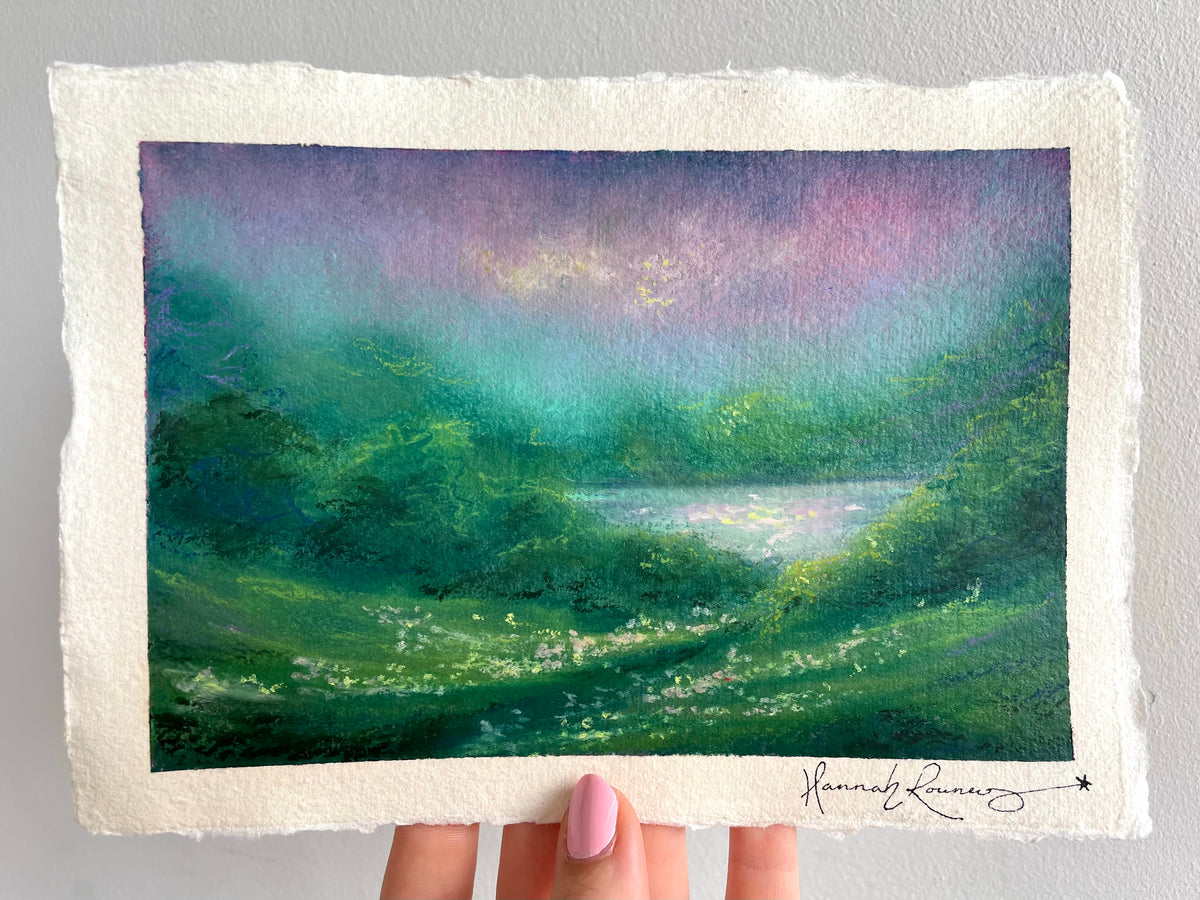 A hand holds a rectangular piece of textured handmade paper featuring an oil pastel painting of a green valley with a lake under a purple and teal sky. The artist's signature, 'Hannah Rowney', is visible in the bottom right corner.