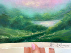 A close-up of a painted landscape artwork showing rolling green hills and a soft pastel sky. The artist signature 'Hannah Rowney' is visible along the bottom edge.