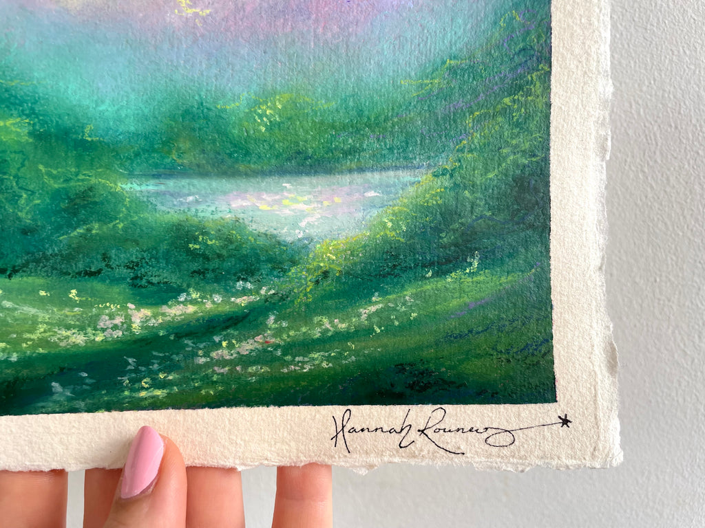 A close-up of a hand holding a landscape painting on textured cream paper. The painting depicts a lush green field with a reflective pond under a soft pastel sky.