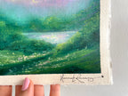 A close-up of a hand holding a landscape painting on textured cream paper. The painting depicts a lush green field with a reflective pond under a soft pastel sky.
