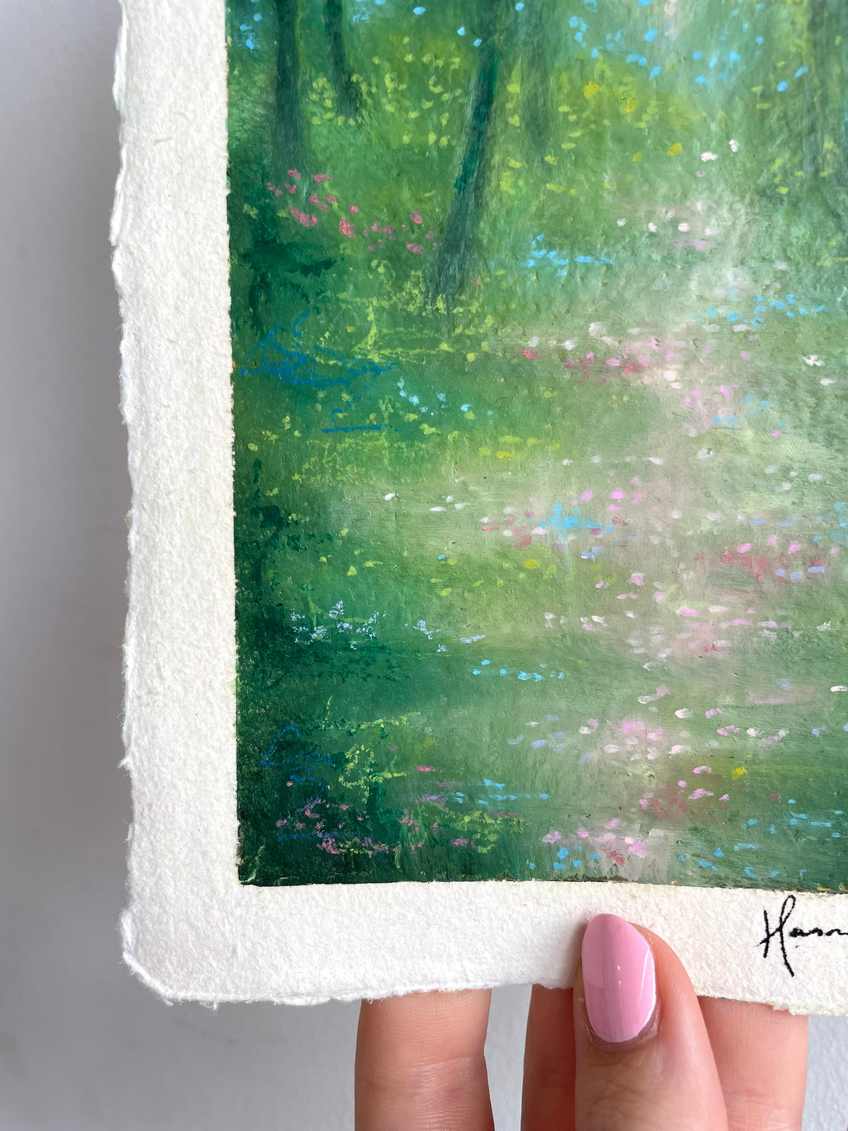 A close-up of a hand holding a vertical oil pastel painting of a forest. The painting features textured green foliage, a soft white path, and small pink and blue floral accents.