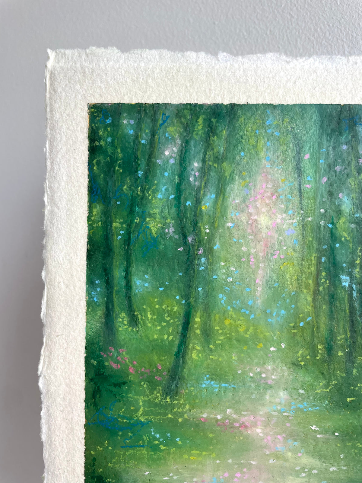A close-up of an oil pastel painting on textured white paper, showing a forest scene with green trees, a soft light beam, and small pink and blue floral accents.